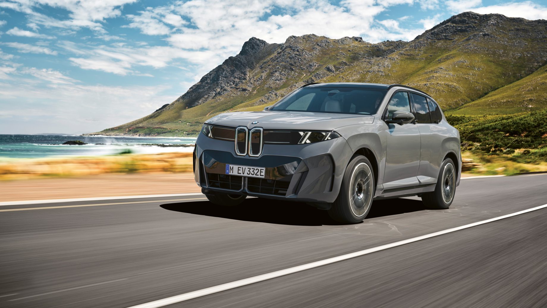 Ein grauer BMW iX Elektro-SUV fährt auf einer Küstenstraße, im Hintergrund sind Berge zu sehen.