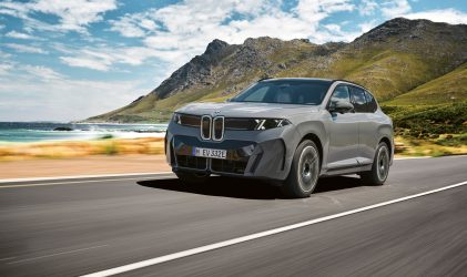 Ein grauer BMW iX Elektro-SUV fährt auf einer Küstenstraße, im Hintergrund sind Berge zu sehen.