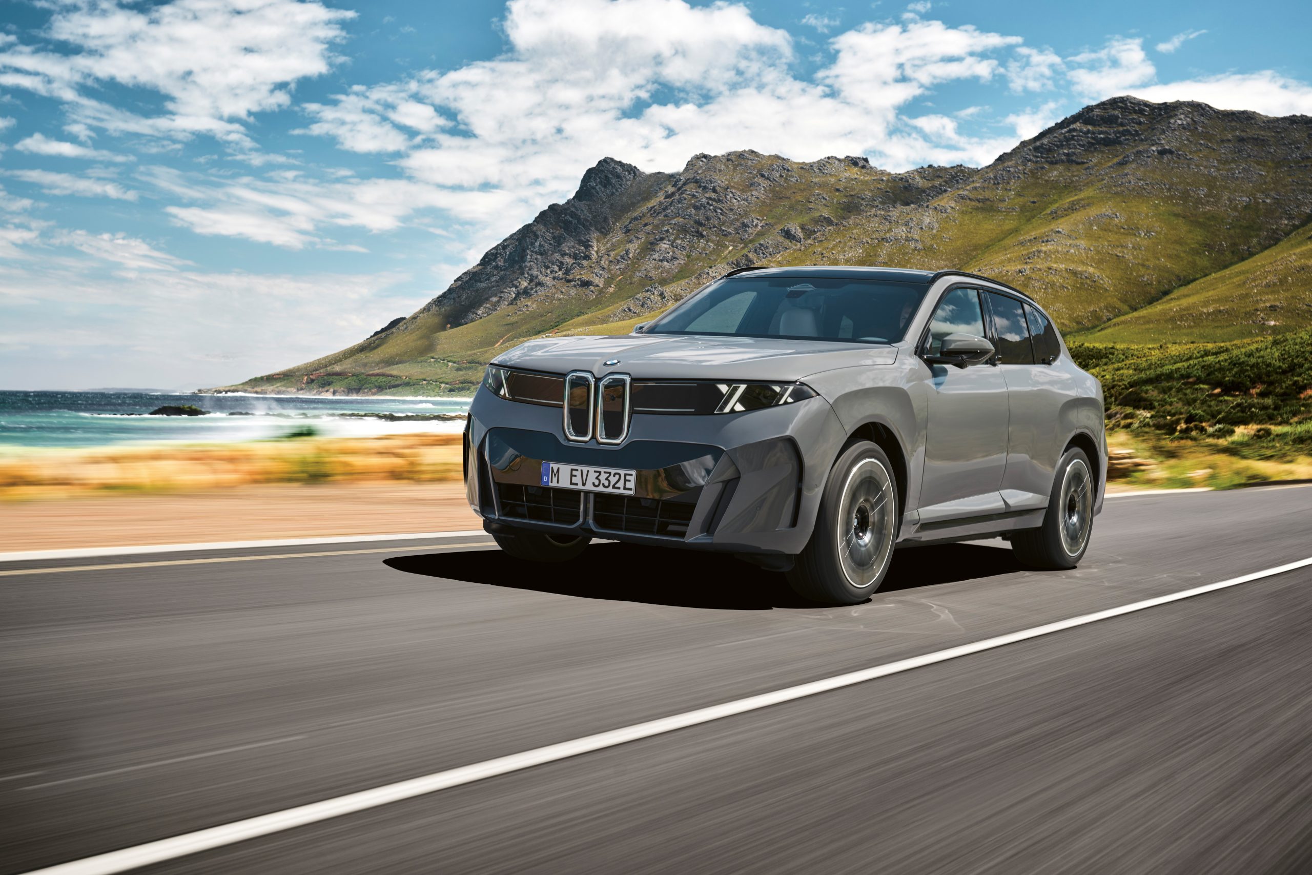 Ein grauer BMW iX Elektro-SUV fährt auf einer Küstenstraße, im Hintergrund sind Berge zu sehen.