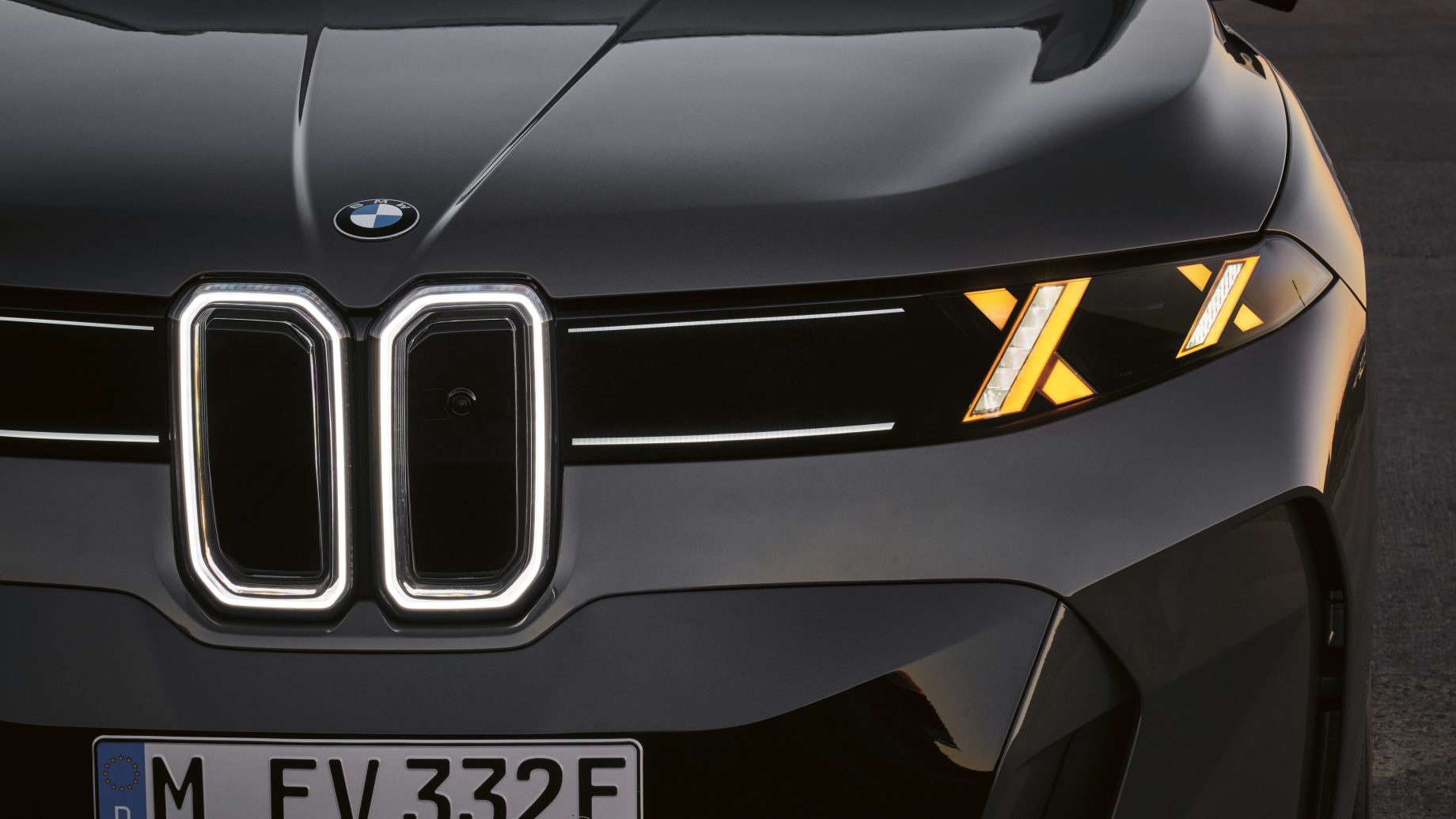 Vorderansicht eines dunklen BMW-Elektroautos mit beleuchteten Konturen des Nierengrills, eckigen, bernsteinfarbenen Tagfahrleuchten und einem deutschen Kennzeichen.