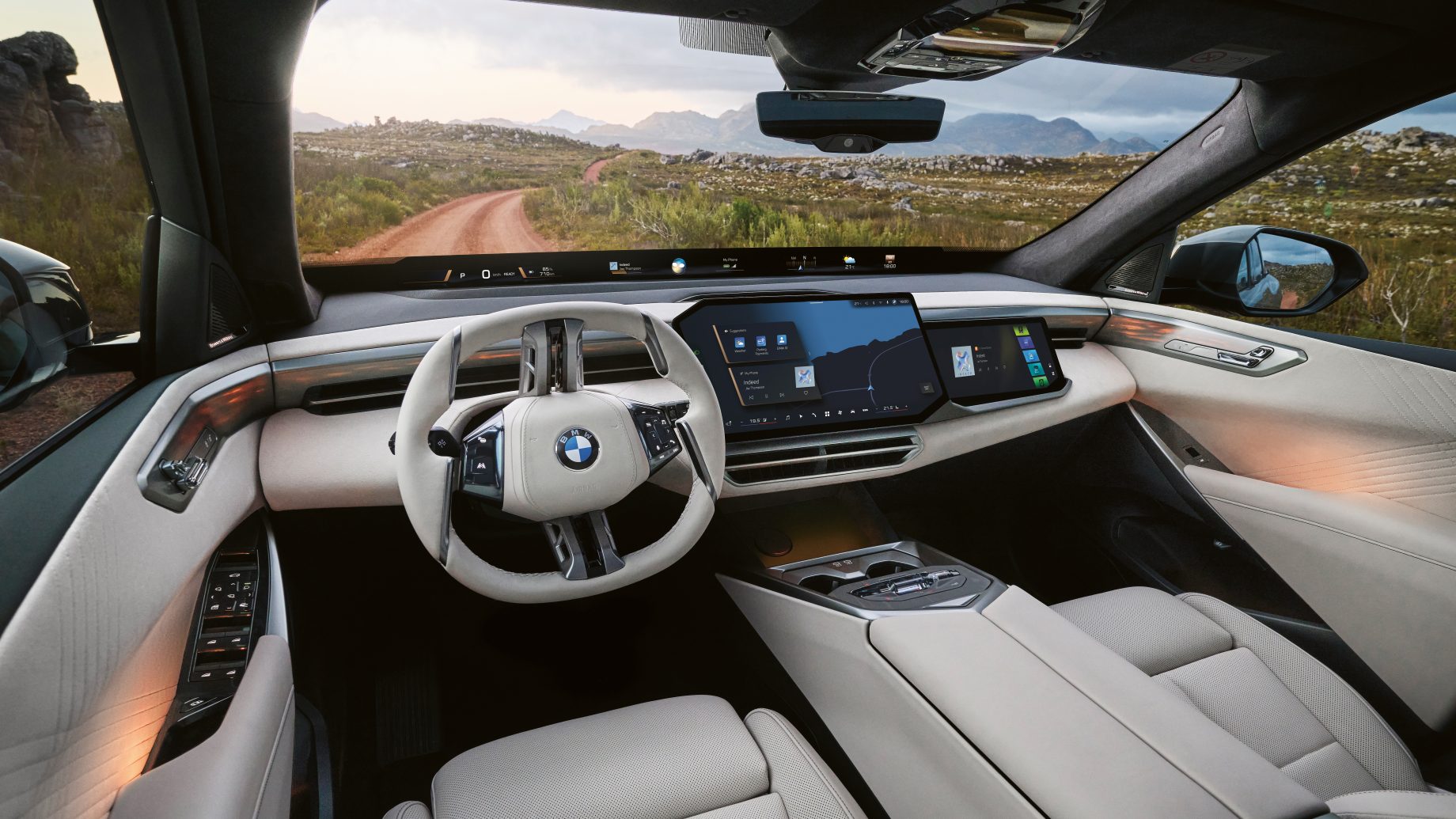 Innenansicht eines BMW mit flachem Lenkrad und großem digitalem Armaturenbrett, mit Blick auf eine unbefestigte Straße in einer Wüstenlandschaft.