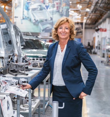 Eine Frau in einem marineblauen Kostüm steht in einer Automobilfabrik neben Autoteilen und Fahrzeugkarosserien auf Montagetischen.