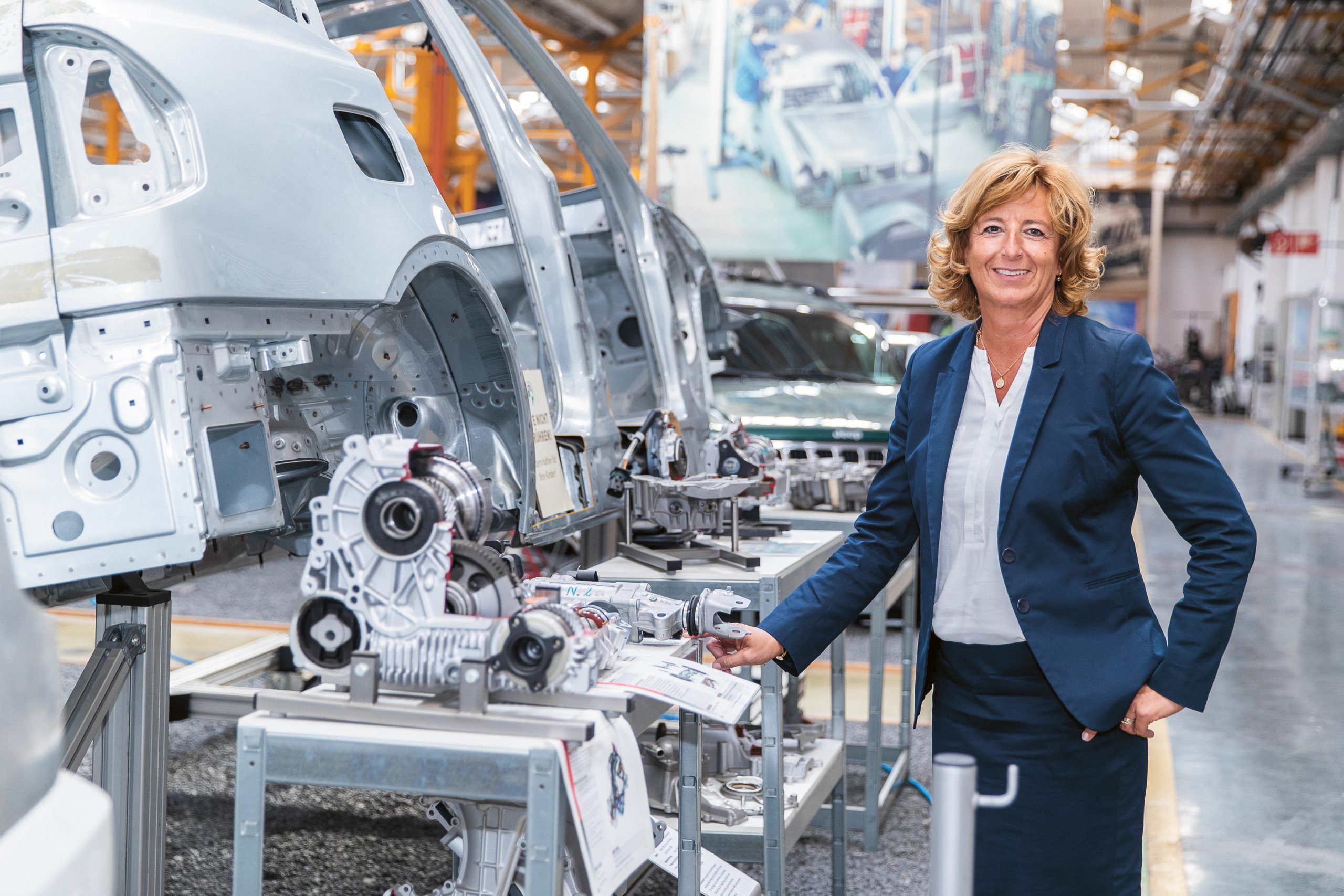 Eine Frau in einem marineblauen Kostüm steht in einer Automobilfabrik neben Autoteilen und Fahrzeugkarosserien auf Montagetischen.