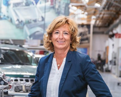 Eine blonde Frau in einem weißen Hemd und einem marineblauen Blazer lächelt, während sie in einer Fabrik oder Werkstatt steht.