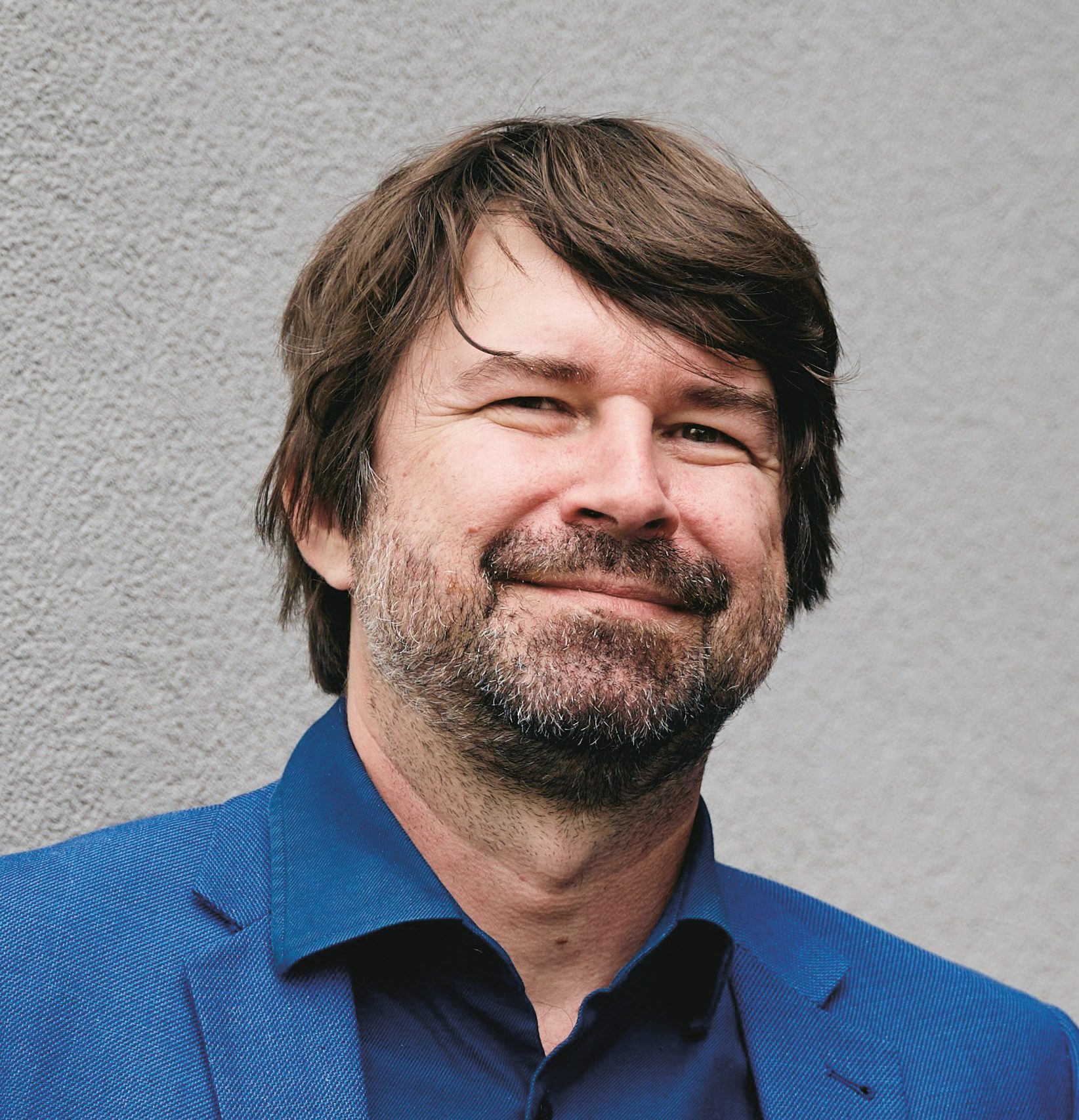 Ein Mann mit braunen Haaren und Bartstoppeln, der ein blaues Hemd mit Knöpfen und einen blauen Blazer trägt.