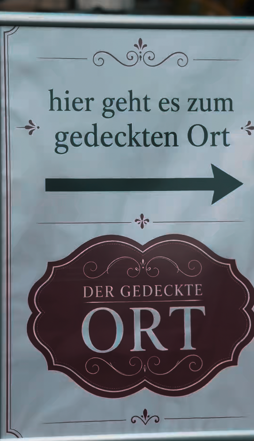 Schild mit einem nach rechts zeigenden Pfeil und der Aufschrift „Hier geht es zum gedeckten Ort“ sowie „DER GEDECKTE ORT“ – ein Millionendeal als Wachstumsturbo.