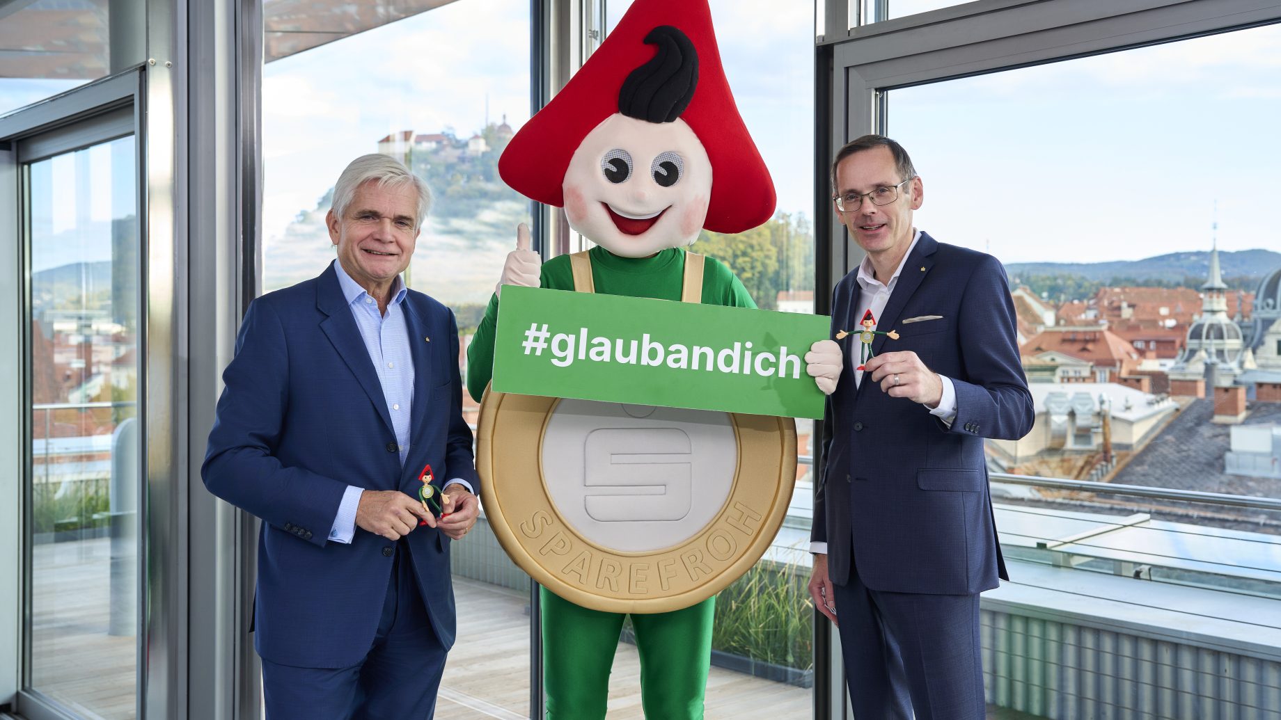 Zwei Männer in Anzügen stehen neben dem Sparefroh-Maskottchen und halten ein Schild von Klampfer Druck mit der Aufschrift „#glaubandich“ in den Händen.