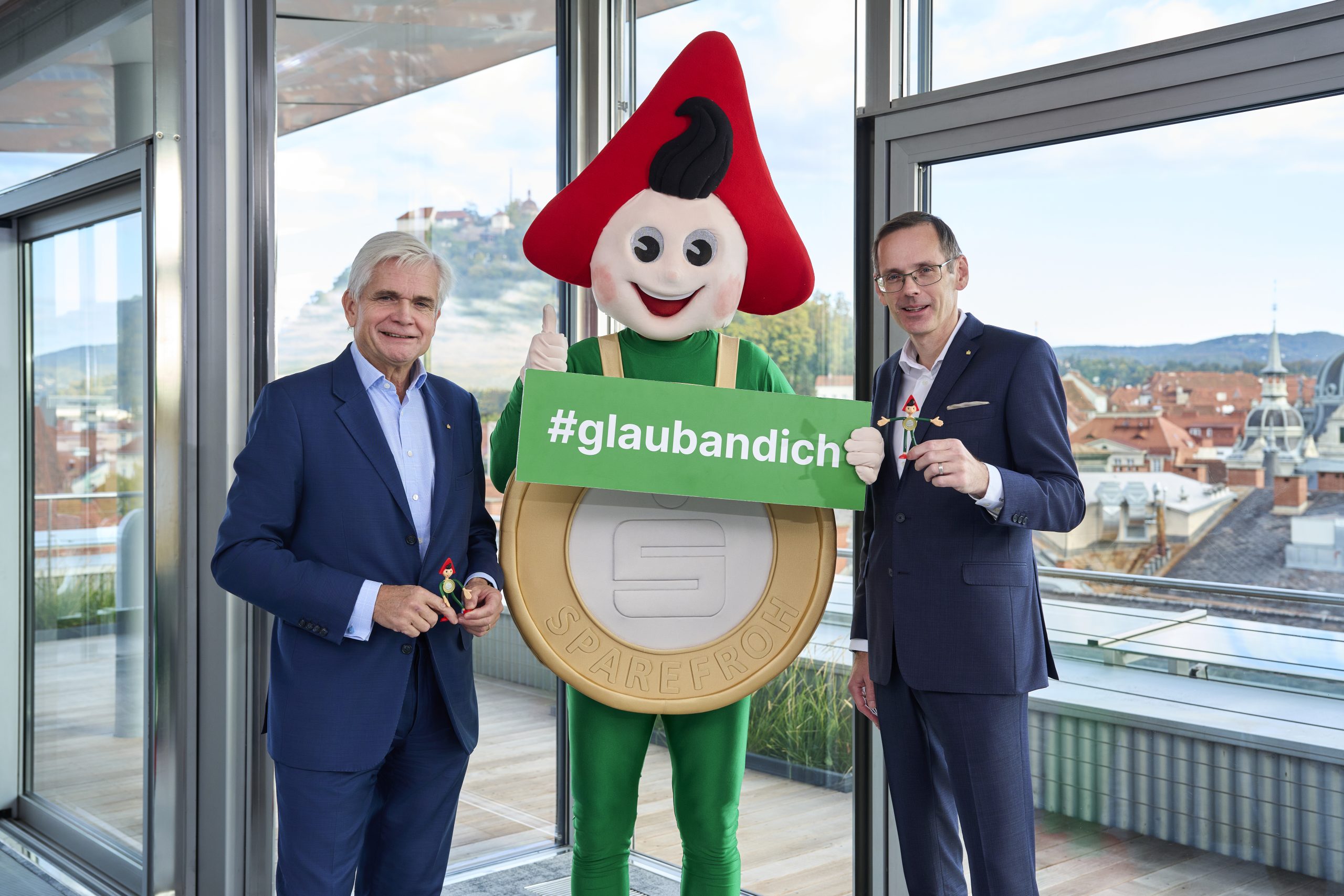 Zwei Männer in Anzügen stehen neben dem Sparefroh-Maskottchen und halten ein Schild von Klampfer Druck mit der Aufschrift „#glaubandich“ in den Händen.