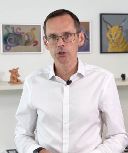 Ein Mann in einem weißen Hemd und mit Brille spricht, während er vor einem Regal mit Kunstwerken steht.