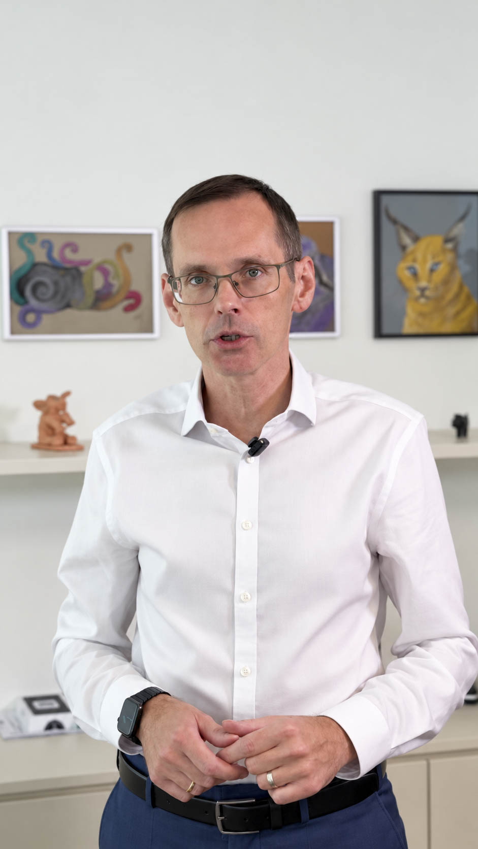 Ein Mann in einem weißen Hemd und mit Brille spricht, während er vor einem Regal mit Kunstwerken steht.