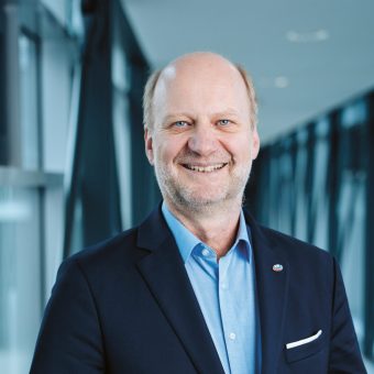 Ein lächelnder Mann mittleren Alters in einem blauen Hemd und einem marineblauen Blazer, der in einem Flur steht.