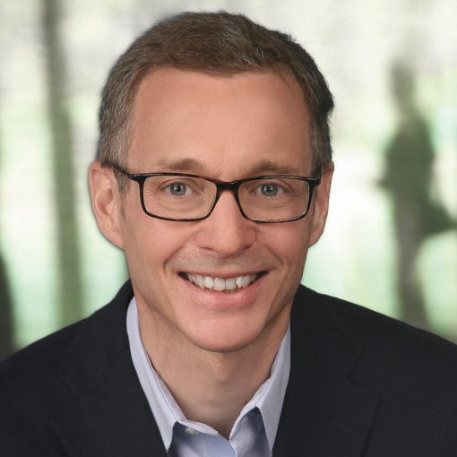 Ein lächelnder Mann mit kurzen braunen Haaren und Brille, der einen dunklen Blazer und ein helles Hemd trägt, vor einem unscharfen Hintergrund.