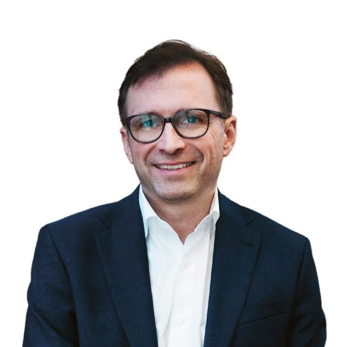 Ein lächelnder Mann mit Brille, einem weißen Hemd mit Kragen und einem dunklen Blazer, der sitzt.