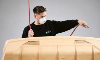 Ein Mann, der eine Schutzmaske trägt, bearbeitet ein großes, speziell geformtes Holzteil mit einem Luftgebläse.