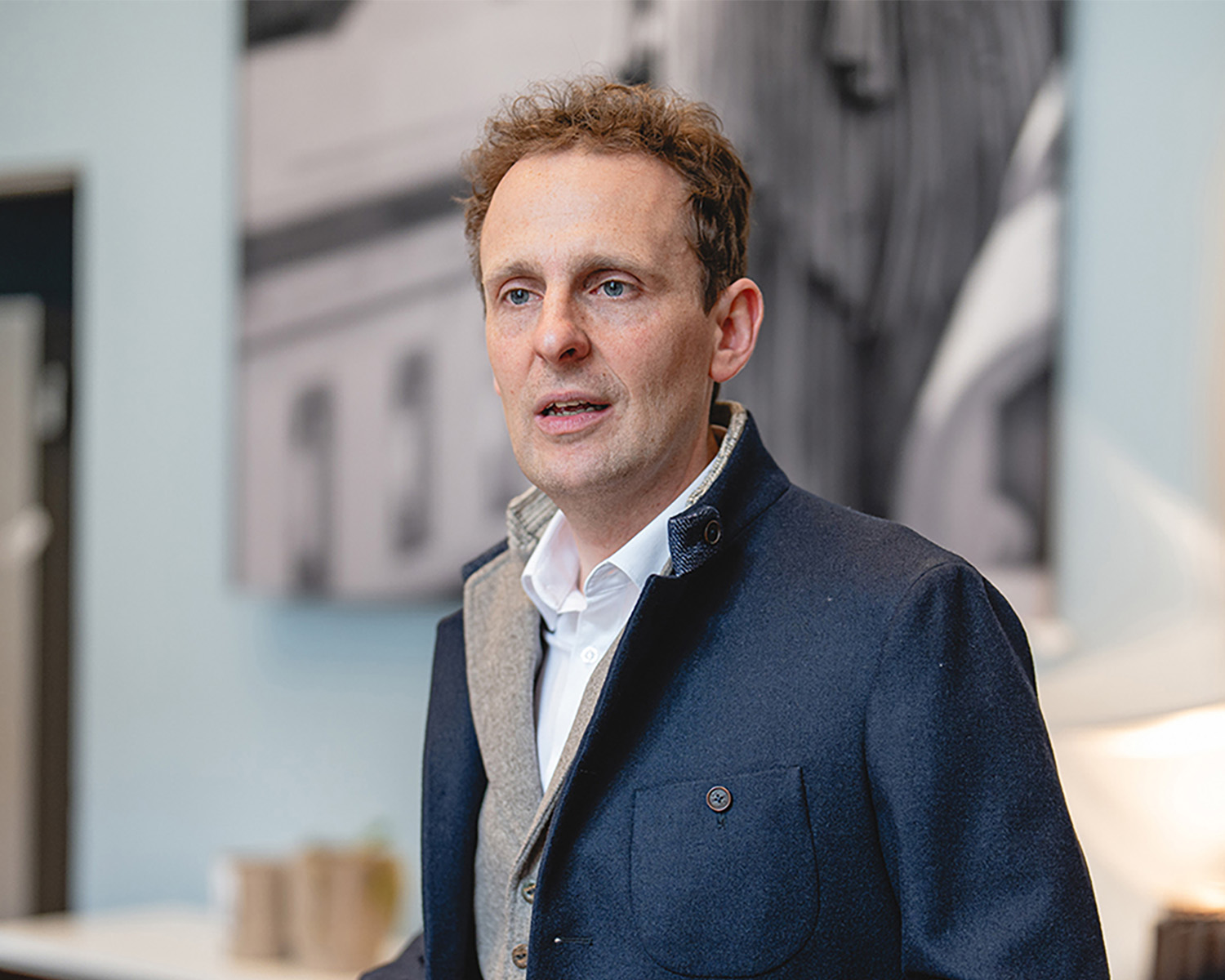 Ein Mann mit kurzen lockigen Haaren, dunkelblauer Jacke und weißem Hemd, steht in einem Haus und spricht oder hört zu. Der Hintergrund ist mit abstrakten Schwarz-Weiß-Kunstwerken an der Wand verschwommen.