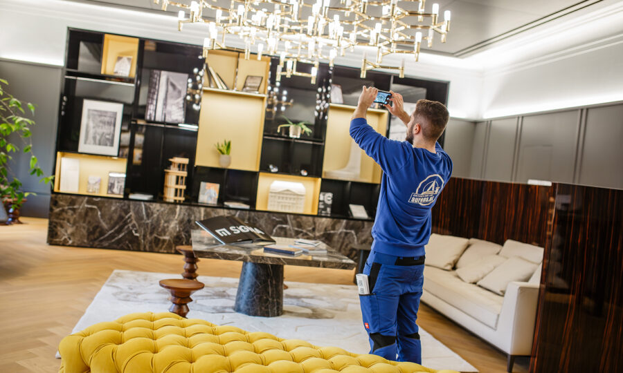 Ein Mann in blauer Arbeitskleidung macht ein Foto mit seinem Handy in einem stilvollen, modernen Wohnzimmer mit einem großen Kronleuchter, einem Marmortisch, einem gelben, getufteten Sofa und Regalen mit Dekorationsartikeln.