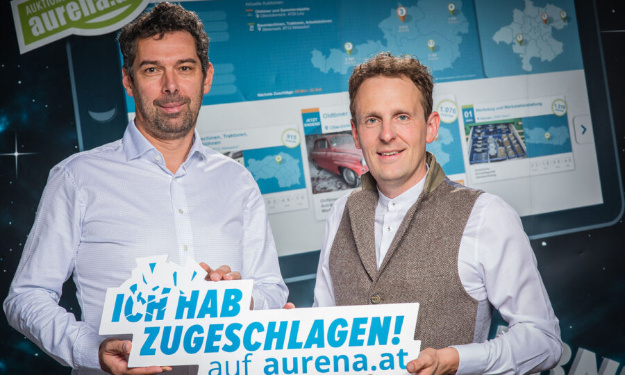 Zwei Männer stehen vor einem digitalen Bildschirm, auf dem Karten und Autos angezeigt werden. Sie halten ein Schild mit der Aufschrift "Ich hab zugeschlagen! auf aurena.at" und lächeln in die Kamera.